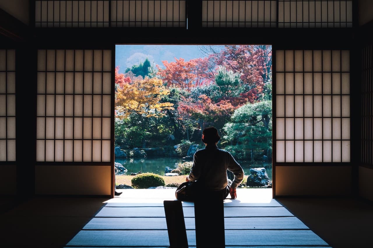 main with hat sat in Japanese house overlooking peaceful pond and yellow/pink cherry blossom trees