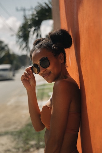 smiling girl with sunglasses against pastel orange wall in sunny place with palm trees in distance